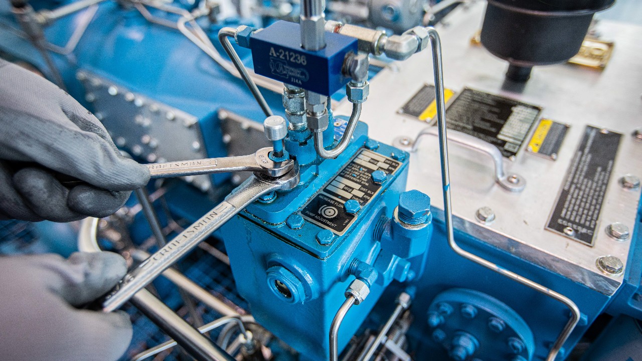 Gloved hands with two wrenches connected to an ariel lube pump, setting the cycle time of the compressor